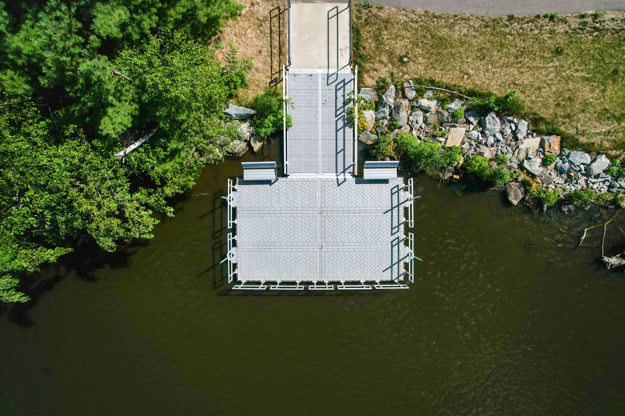 View of a T-shaped, gray, modular floating dock system extending from a grassy and rocky shoreline into dark green water