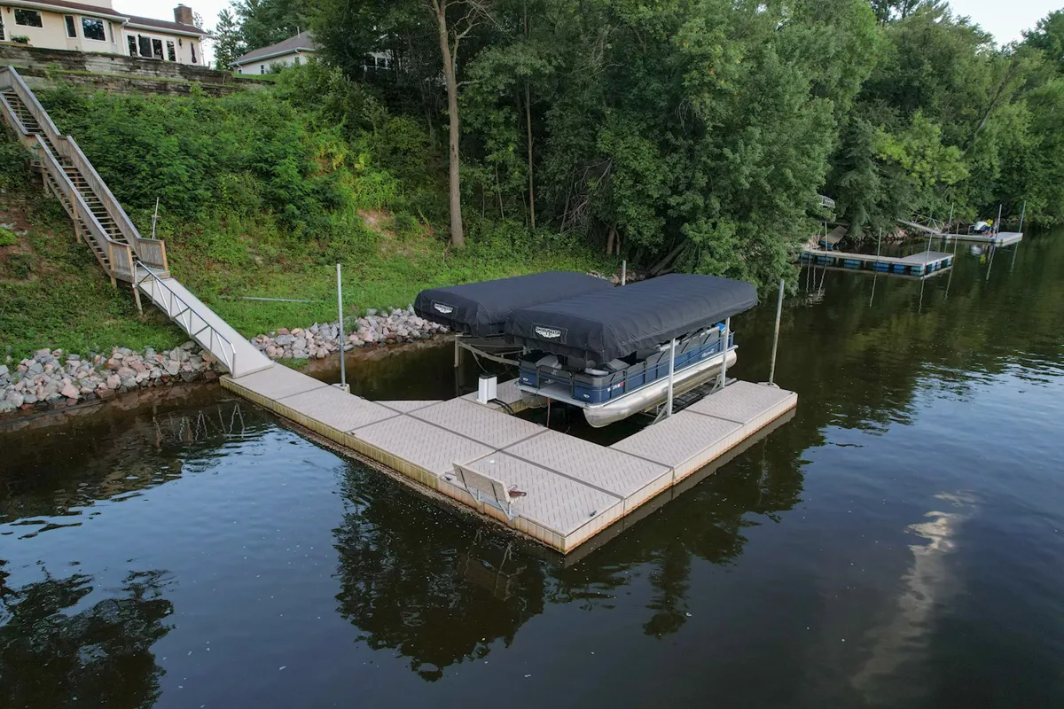 A pontoon boat sits covered under a canopy attached to a modular dock system on a body of water.