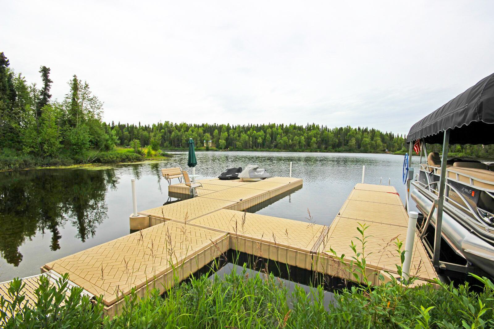 A tan modular floating dock extends into a calm lake, with a covered boat moored to the right and a wooded shoreline in the background. 